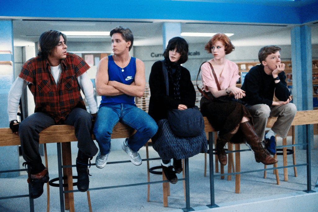 Photo of a scene from the movie The Breakfast Club, with the characters sitting on a library railing. 