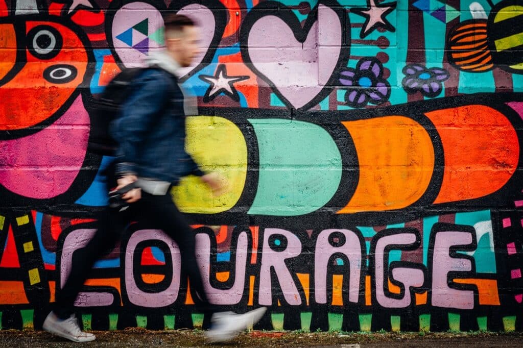 Photo of man with a backpack walking past a colorful wall mural that says Courage