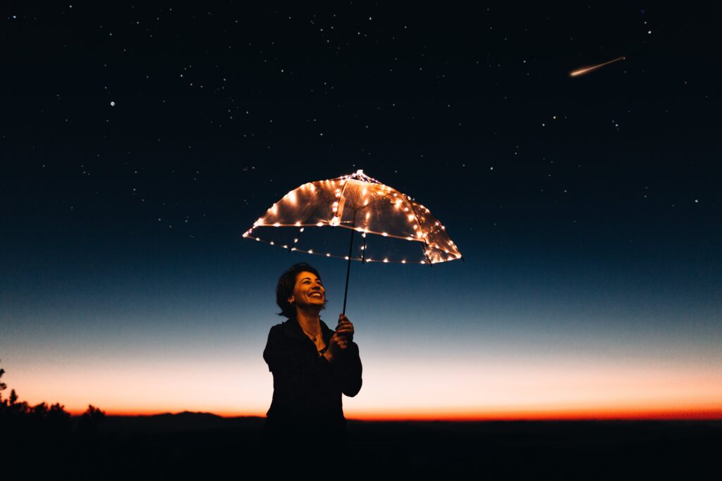 Photograph of a smiling woman at sunset, holding a clear umbrella that is lined with small, bright lights., with stars and a shooting star in the sky.