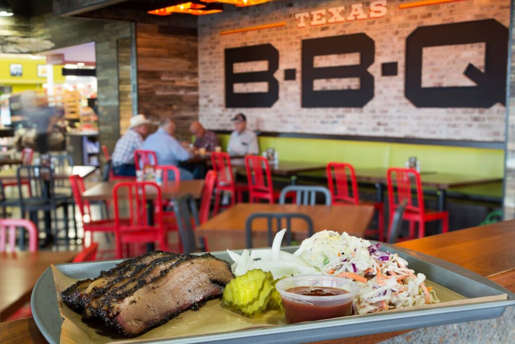 Photograph inside of a BBQ restaurant, highlighting a plate of brisket and coleslaw, with giant letters B B Q on the wall. Grab some classic Texas BBQ while at Claris Engage. 