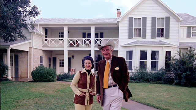 Photograph of Lady Bird and Lyndon Johnson, in front of a white home at the LBJ Ranch near Austin, Texas.