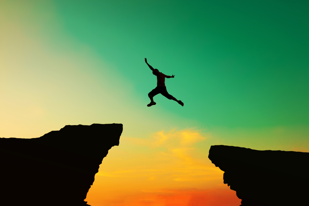Photo of a person jumping between two rock outcroppings, with a vibrant sunrise in the background.