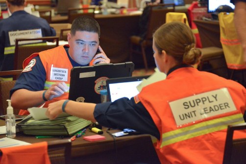 Photo of two people assisting during a disaster response - wearing high visibility orange and yellow vests, labeled with their areas of Logistics and Supply.