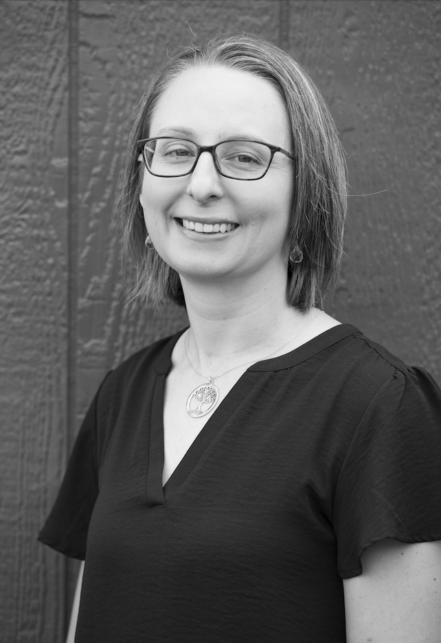 Headshot for Annie Woyna. A white woman in her 30s with shoulder length hair and square, black glasses.