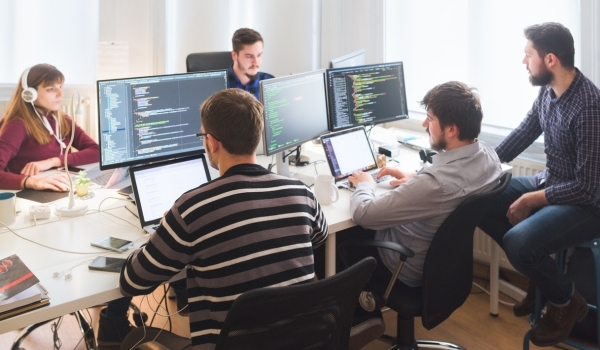 A group of five people sitting at a desk and coding. Three are on the side closest to the camera and the other two are on the opposite side of the desk. The woman on the opposite side and far left is wearing headphones. This article talks about the benefits of using separate development and production environments.