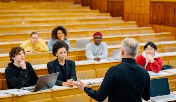 A professor teaching a class to a group of college students. Some have laptops, some have pens and paper. Illustrating a college who upgraded to the latest version of FileMaker.